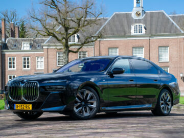 Een strakke, zwarte luxe sedan staat geparkeerd voor een groot bakstenen gebouw met een klok op de gevel. In de BMW i7-recensie wordt beschreven dat hij geavanceerde functies en weelderig comfort heeft, perfect passend bij de scène. Op het gele kenteken staat 'R-152-RG', terwijl bomen het gebouw omlijsten.