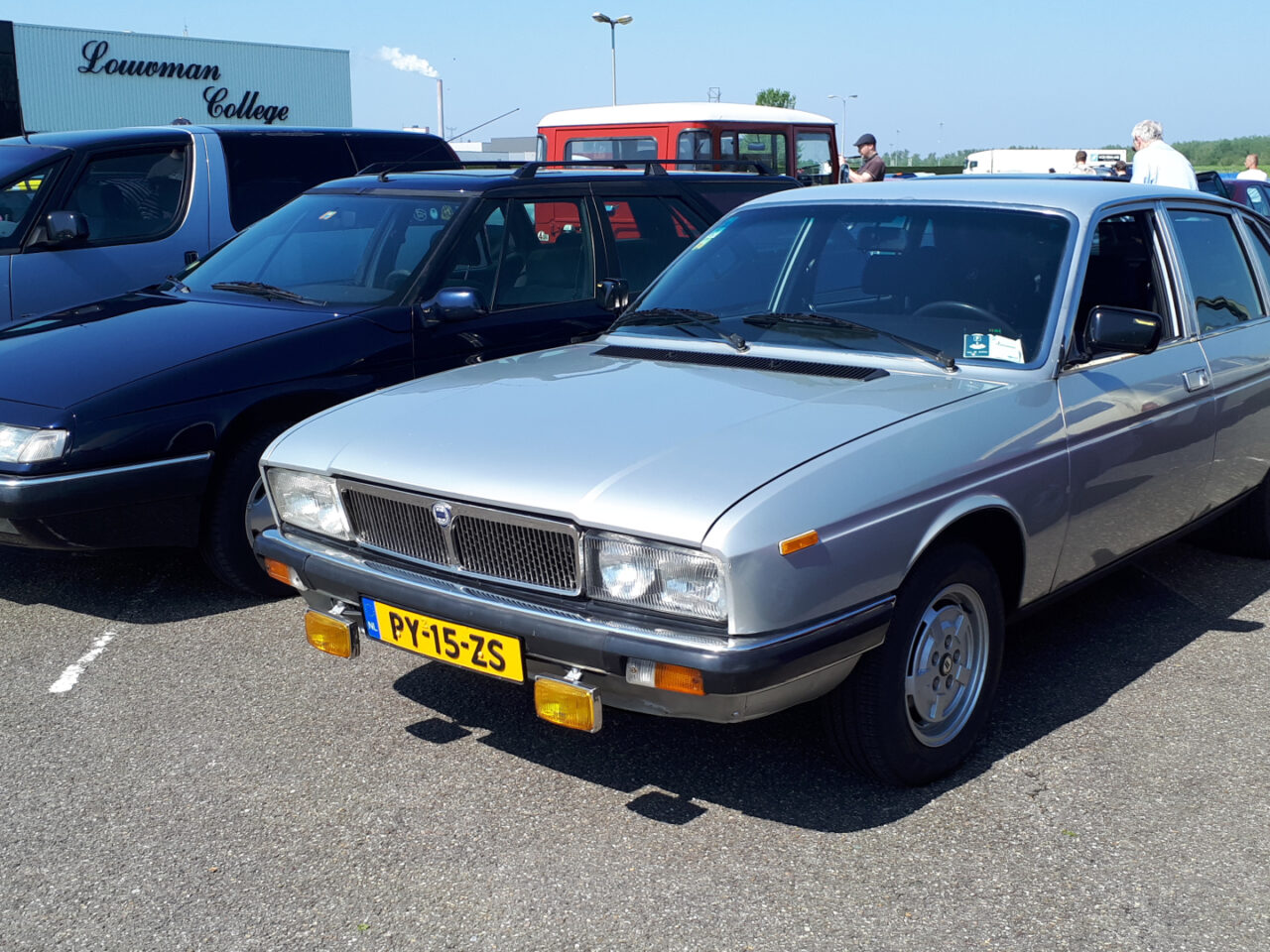Onder een heldere hemel staan verschillende oldtimers geparkeerd. De focus ligt op een zilveren sedan met Nederlandse kentekenplaten voorop, die doet denken aan een tentoonstelling bij EMWalhalla.