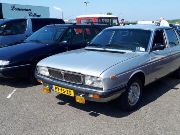 Onder een heldere hemel staan verschillende oldtimers geparkeerd. De focus ligt op een zilveren sedan met Nederlandse kentekenplaten voorop, die doet denken aan een tentoonstelling bij EMWalhalla.