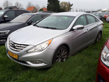 Een zilveren Hyundai Sonata met gele kentekenplaten staat naast andere auto's op het gras geparkeerd.