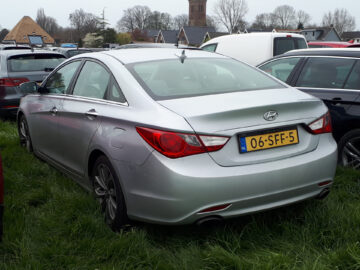 Een zilverkleurige Hyundai Sonata sedan staat tussen andere voertuigen op het gras geparkeerd. De auto heeft een Nederlands kenteken met nummer 06-SFF-5. Op de achtergrond zijn bomen en gedeeltelijke gebouwen zichtbaar.