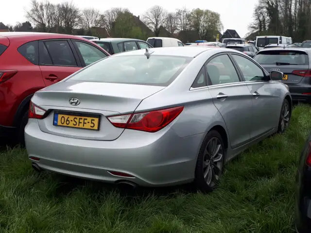 Een zilveren Hyundai Sonata sedan met Nederlands kenteken, geparkeerd op een grasveld omringd door andere voertuigen.