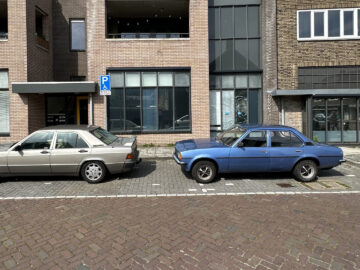 Twee geparkeerde auto's, een beige en een blauwe, voor een bakstenen bedrijfsgebouw. De beige auto staat op een gemarkeerde parkeerplaats en er staat een bord met de parkeerregels. Het is alsof je in de tijd teruggaat naar een eenvoudiger tijdperk met klassieke voertuigen en tijdloze architectuur.