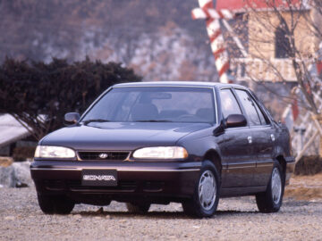 Een donkere Hyundai Sonata sedan geparkeerd op een grindoppervlak voor bomen en een gebouw. Op het kenteken staat "SONATA.