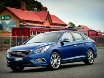 Een blauwe Hyundai Sonata sedan staat geparkeerd voor een historisch treinstation met een rood dak en een vintage trein op de baan.