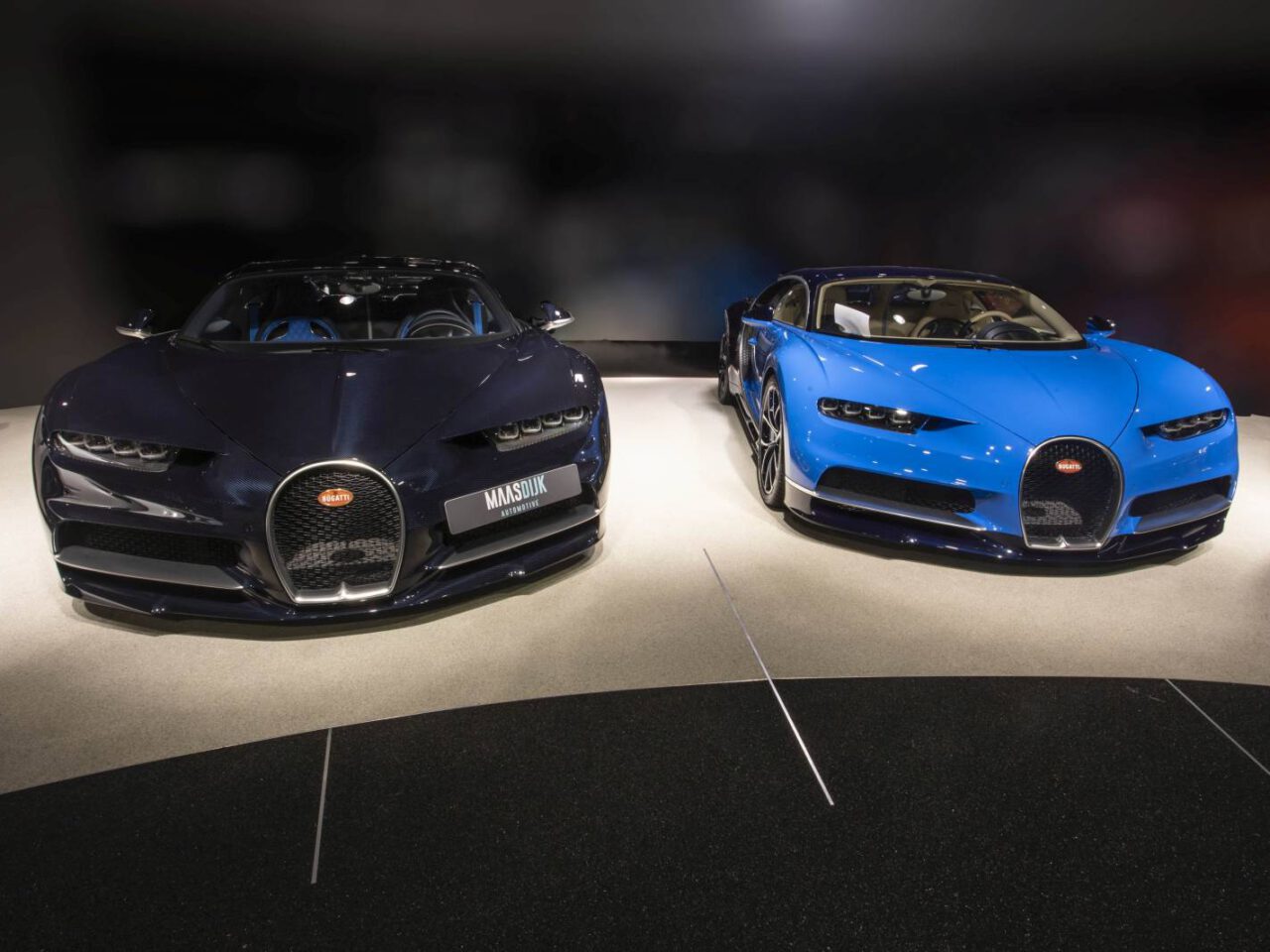 Twee Bugatti Chiron-sportwagens worden naast elkaar tentoongesteld, één in het zwart en de andere in blauw, op een showroomvloer.