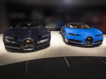 Twee Bugatti Chiron-sportwagens worden naast elkaar tentoongesteld, één in het zwart en de andere in blauw, op een showroomvloer.