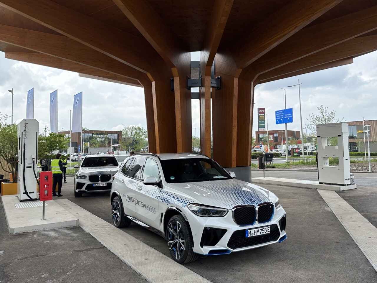 Twee witte BMW-voertuigen staan bij een waterstoftankstation en laten de mogelijkheden van rijden op waterstof zien. De ene auto wordt door iemand bijgetankt terwijl de ander wacht. De houten dakconstructie en uitrusting zijn zichtbaar.