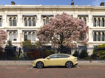 Een gele auto, mogelijk een Nio ET5, staat geparkeerd in een geplaveide straat voor een wit gebouw met boogramen en een bloeiende boom.