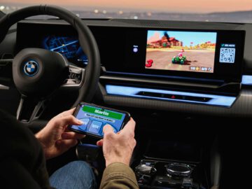 Persoon in een BMW i5 die een racespel speelt op een mobiele telefoon, terwijl het dashboard van de auto hetzelfde spel op een groter scherm weergeeft.