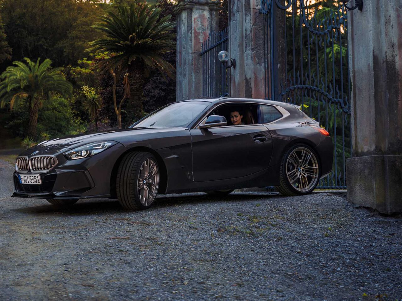 Een donkergrijze BMW Concept Touring Coupé met daarin een persoon staat geparkeerd bij een sierlijk smeedijzeren hek met stenen pilaren, tegen een achtergrond van weelderig groen en palmbomen.