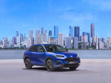 De strakke blauwe Honda ZR-V SUV staat geparkeerd voor de skyline van een stad, met hoge gebouwen en een helderblauwe lucht die een adembenemende achtergrond vormen.