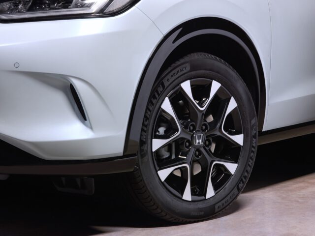 Close-up van het voorwiel van een witte Honda ZR-V met een Michelin-band en een zwart-zilveren lichtmetalen velg, waarbij een deel van de koplamp en bumper van de auto zichtbaar is.