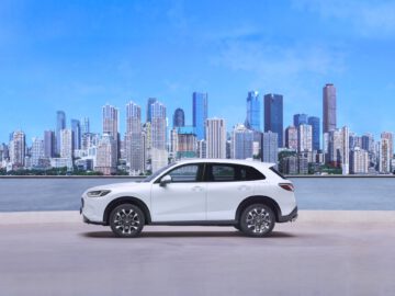 Een witte Honda ZR-V SUV staat geparkeerd op een verhard oppervlak met de skyline van de stad en een watermassa op de achtergrond. De lucht is helder en blauw.