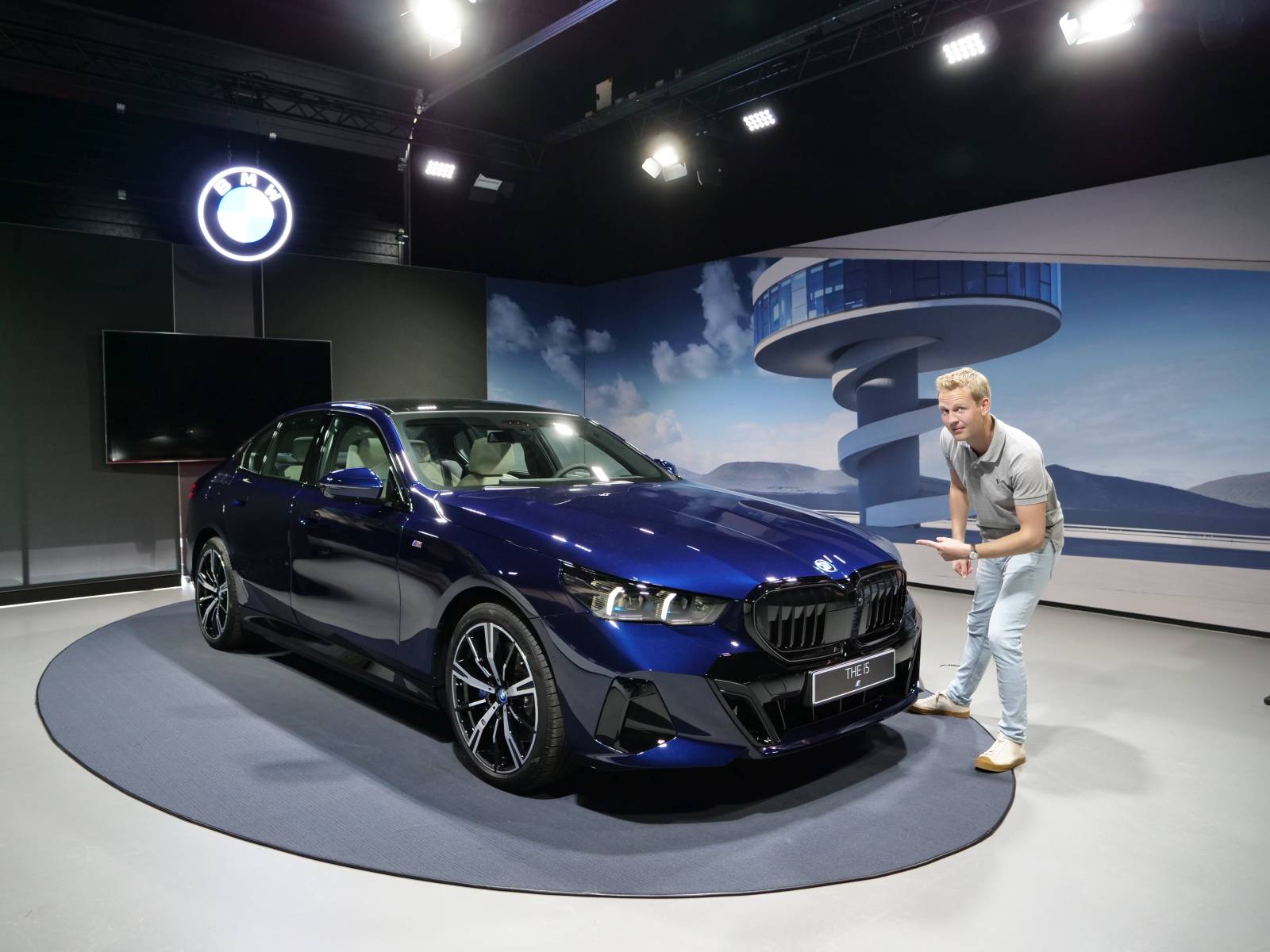 Een persoon staat naast een strakke blauwe BMW i5 in een showroom, gemarkeerd door een prominent BMW-bord en een groot scherm met een futuristisch gebouw op de achtergrond.