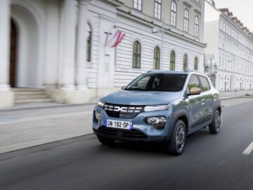Een blauwe Dacia Spring SUV rijdt door een stadsstraat met witte gebouwen, waarvan er één een rood-wit spandoek heeft. Op het kenteken staat "GN-182-DP".