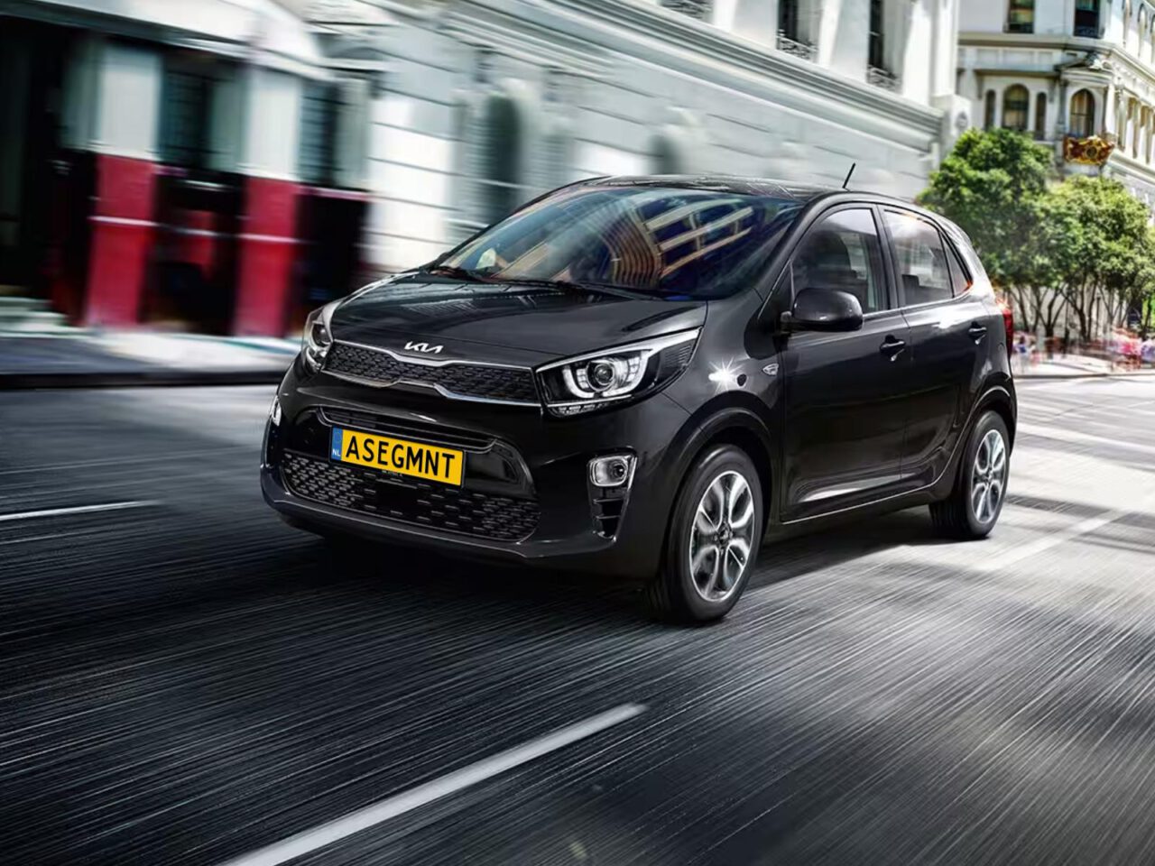 Een zwarte compacte auto uit het A-segment met kenteken "ASEGMNT" rijdt door een stadsstraat omzoomd met gebouwen en bomen.