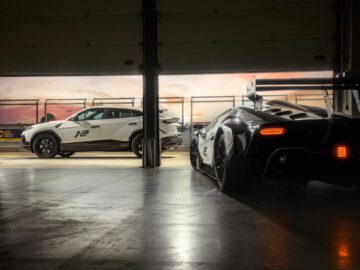 Twee sportwagens, een zwarte en een witte, staan geparkeerd in een garage die bij zonsondergang uitkomt op een racecircuit. De zwarte auto staat op de voorgrond en kijkt weg, terwijl de witte Lamborghini Urus SC-V12 Edition net buiten de garage staat.
