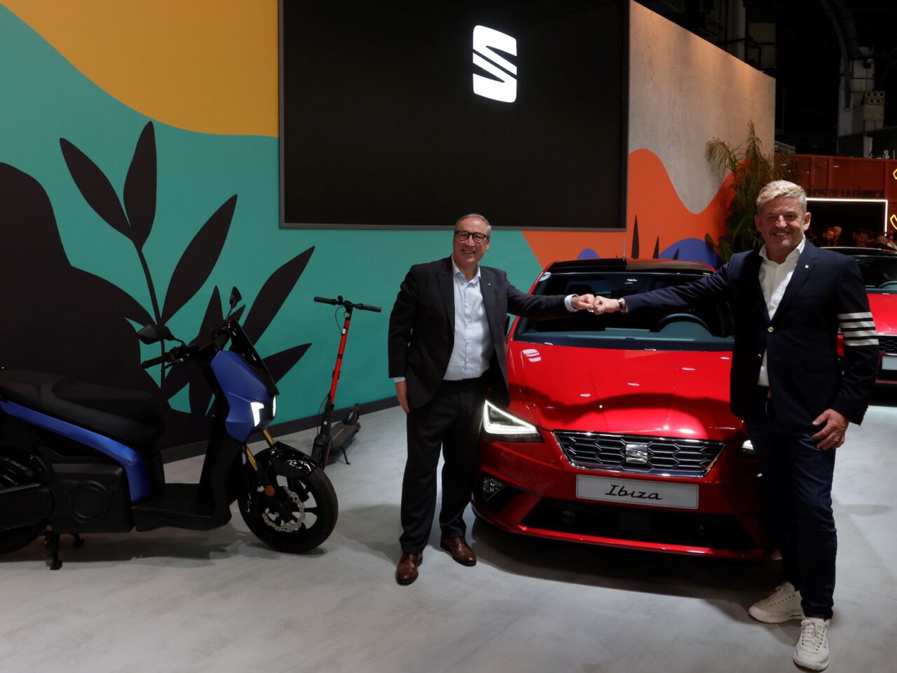 Twee mannen in formele kledij staan voor een rode SEAT Ibiza-auto elkaar de hand te schudden. Naast hen staan een elektrische scooter en een ander voertuig. Op de achtergrond is een kleurrijke muur met het prominente SEAT-logo zichtbaar.