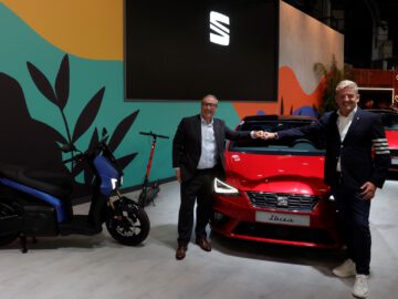 Twee mannen in formele kledij staan voor een rode SEAT Ibiza-auto elkaar de hand te schudden. Naast hen staan een elektrische scooter en een ander voertuig. Op de achtergrond is een kleurrijke muur met het prominente SEAT-logo zichtbaar.