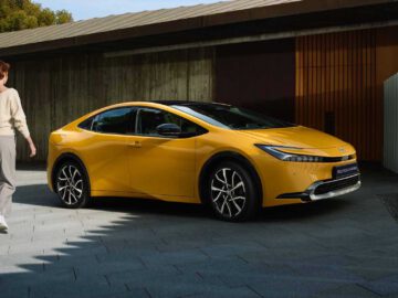 Een persoon in vrijetijdskleding loopt naar een gele Toyota Prius 2023, geparkeerd op een moderne oprit met een gebouw op de achtergrond.