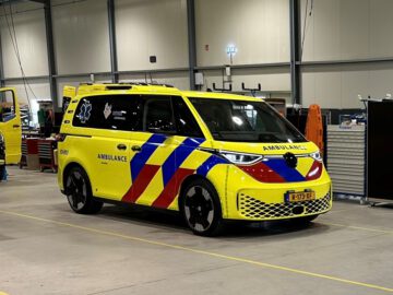Een geel-rood gestreepte Volkswagen ID.Buzz ambulancebusje staat geparkeerd in een ruime, goed verlichte garage met gereedschap en uitrusting op de achtergrond.