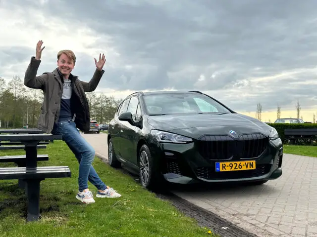 Een persoon steekt opgewonden zijn handen op terwijl hij op een bankje staat naast een geparkeerde zwarte BMW 230e Active Tourer met een geel kenteken. Bomen en een bewolkte hemel zijn op de achtergrond.