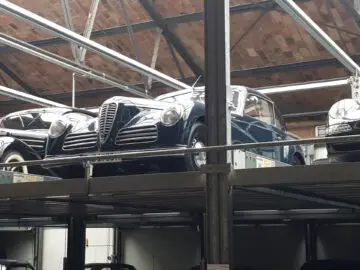 Een klassieke auto staat geparkeerd op een verhoogd platform in Classic Remise, een pakhuis met een hoog bakstenen plafond. Eromheen zijn andere oldtimers zichtbaar.