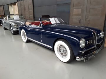 Bij Classic Remise staan twee vintage cabrio's geparkeerd. De auto op de voorgrond is donkerblauw met witwandige banden, terwijl de auto erachter straalt van zilveren elegantie.