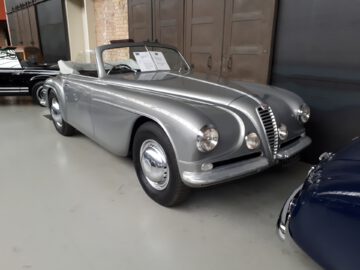 Een vintage zilveren cabriolet staat binnen geparkeerd bij Classic Remise, met informatieborden op de voorruit.