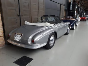 Bij Classic Remise staat een grijze vintage cabriolet binnen geparkeerd naast andere klassieke auto's. Op het kenteken staat "RONY 124.