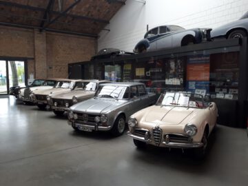 Bij Classic Remise wordt binnen een collectie oldtimers tentoongesteld. Vier klassieke auto's staan op een rij onder een tussenverdieping, waar een andere auto boven staat. Op de achtergrond zijn borden en posters zichtbaar.