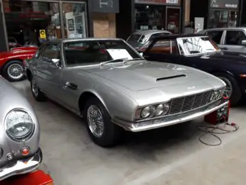 Een zilveren klassieke auto met een prominente grille en spaakwielen wordt tentoongesteld in een Classic Remise-showroom, omringd door andere vintage voertuigen. Op de voorruit wordt een bord geplaatst.