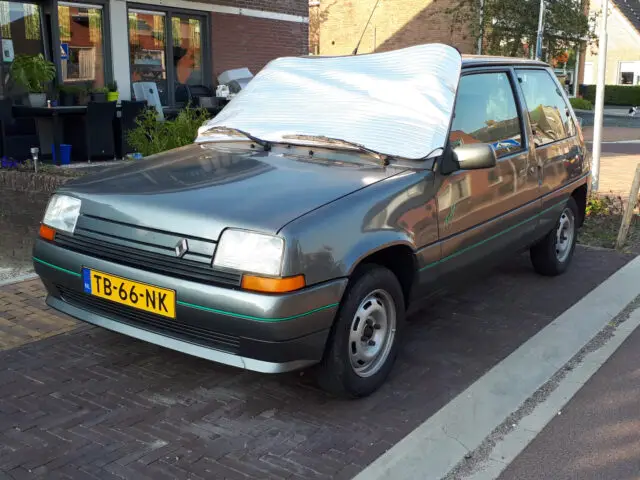 Op straat staat een grijze Renault 5 geparkeerd met een reflecterend zonnescherm over de voorruit. Het voertuig heeft gele en zwarte kentekenplaten en staat naast een met stenen geplaveid trottoir.