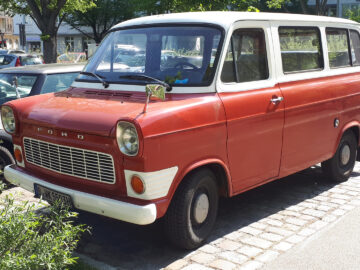 Een rood-wit vintage Ford-busje, recentelijk gespot, staat geparkeerd in een geplaveide straat vol met andere voertuigen. Op de achtergrond zijn bomen en gebouwen zichtbaar.