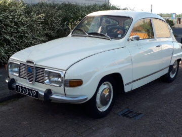 Een witte klassieke Saab 96 staat geparkeerd op een verhard terrein nabij struiken. Op het kenteken staat "11-20-VP.