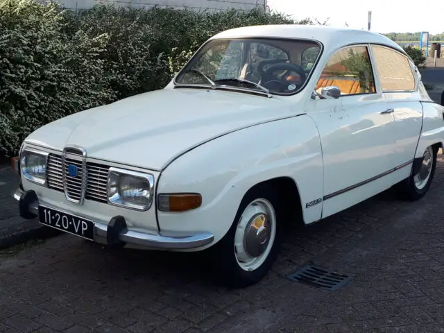 Een witte klassieke Saab 96 staat geparkeerd op een verhard terrein nabij struiken. Op het kenteken staat 