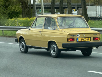 Een gele vintage Volvo 66 met kenteken "16-VZ-67" rijdt op een snelweg grenzend aan een groenstrook.