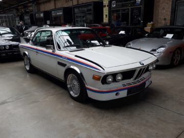 Bij Classic Remise staat onder andere een witte BMW 3.0 CSL met kleurrijke racestrepen binnen geparkeerd. Het voertuig heeft een vintage ontwerp met luchtinlaten aan de voorkant, strakke lijnen en verchroomde wielen.