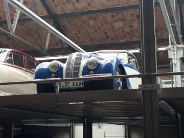 Een blauwe vintage auto staat geparkeerd op een metalen platform in een gebouw met een houten plafond, dat doet denken aan de klassieke sfeer die je bij Classic Remise aantreft.