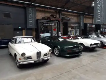 Verschillende klassieke auto's, waaronder een witte en donkergroene, worden tentoongesteld in een ruime overdekte showroom bij Classic Remise, met 'Thiesen'-banners die aan het plafond hangen.