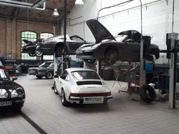 Een autoservicegarage bij Classic Remise met verschillende sportwagens op liften en monteurs hard aan het werk. Twee auto's staan verhoogd met de motorkap open, terwijl drie andere op de grond staan, waaronder een witte sportwagen op de voorgrond.