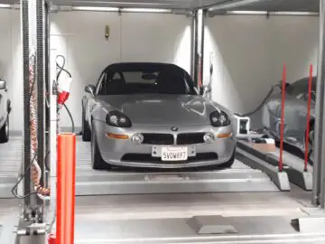 Een zilveren auto met een Californisch kenteken staat geparkeerd op een autolift met meerdere niveaus in een op Classic Remise geïnspireerde garage.