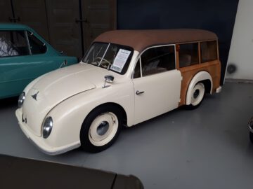 Crèmekleurige vintage auto met een bruin softtopdak en houten achterpanelen, binnen tentoongesteld bij Classic Remise. Op het raam aan de bestuurderszijde is een briefje bevestigd.