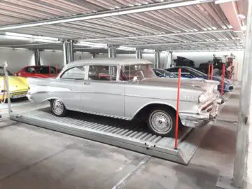 Een klassieke grijze tweedeursauto staat geparkeerd in een autostalling met meerdere verdiepingen bij de gerenommeerde Classic Remise, omringd door andere voertuigen.