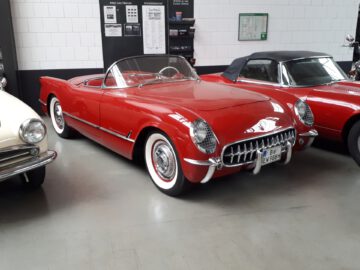 Een klassieke rode cabriolet met whitewall-banden staat binnen geparkeerd bij Classic Remise, omringd door andere oldtimers. Op het kenteken staat "EV 5884.