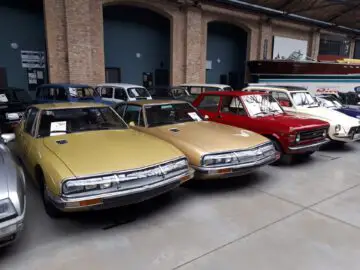 Een verzameling oldtimers die binnen bij Classic Remise wordt tentoongesteld, waaronder gele, bruine en rode voertuigen. Eén auto heeft een zichtbaar veilingbord op de voorruit. De achtergrond is voorzien van een boot en bakstenen muren.