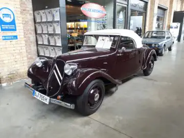 Een kastanjebruine vintage auto met een wit dak wordt tentoongesteld in een garage bij Classic Remise. Op de achtergrond zijn borden en een andere oldtimer zichtbaar. De auto heeft een displaycard op de voorruit en een Duits kenteken.