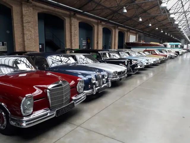 In de overdekte Classic Remise showroom met een hoog plafond in industriële stijl staat een rij klassieke auto’s tentoongesteld. De auto's staan geparkeerd naast een bakstenen muur met gebogen deuropeningen.