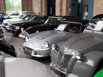 Een rij oldtimers die binnen worden tentoongesteld bij Classic Remise; de voertuigen variëren in kleur, merk en model, met informatieve borden op de voorruit.